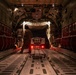 The image shows the interior of a large military cargo aircraft, likely a C-17 Globemaster or C-5 Galaxy, with the rear ramp open. Personnel in military flight suits are overseeing the loading or unloading of a military vehicle or weapons system via the rear cargo ramp at night. The cargo hold is illuminated by interior lighting, with red tail lights from the vehicle visible near the ramp opening.