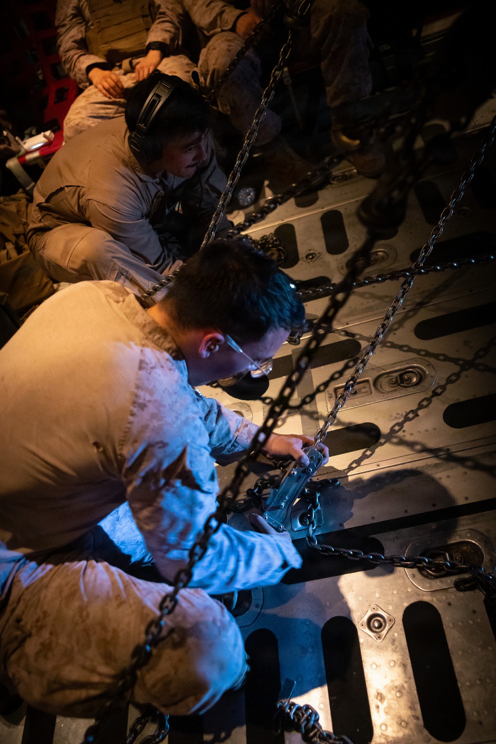 Military personnel in camouflage uniforms are working inside what appears to be a military transport aircraft cargo hold. The foreground individual is actively securing or adjusting heavy chains and tie-down equipment to the aircraft floor. Additional personnel are visible seated in the background of the dimly lit cargo bay.