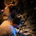 Military personnel in camouflage uniforms are working inside what appears to be a military transport aircraft cargo hold. The foreground individual is actively securing or adjusting heavy chains and tie-down equipment to the aircraft floor. Additional personnel are visible seated in the background of the dimly lit cargo bay.