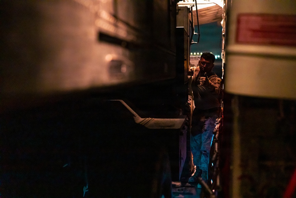 A person is standing in a narrow gap between two large vehicles at night, illuminated by ambient artificial lighting. The scene appears to be an industrial or transportation setting, possibly a loading dock, depot, or roadside stop. The low-light conditions and tight framing create a dramatic, shadowy composition.