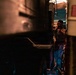 A person is standing in a narrow gap between two large vehicles at night, illuminated by ambient artificial lighting. The scene appears to be an industrial or transportation setting, possibly a loading dock, depot, or roadside stop. The low-light conditions and tight framing create a dramatic, shadowy composition.
