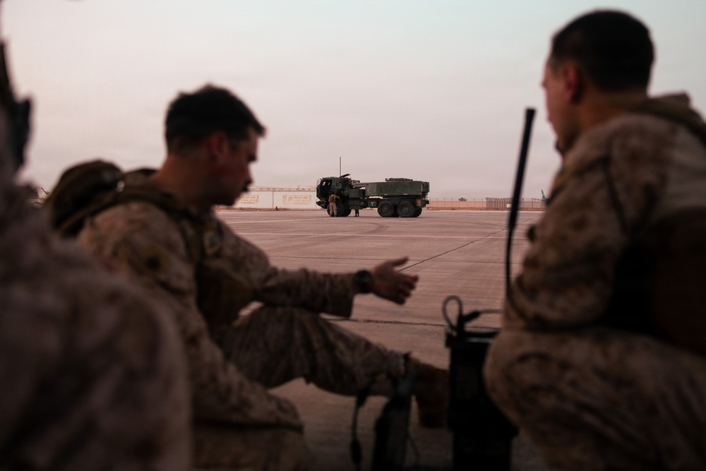 Two military personnel in desert camouflage uniforms are observed in the foreground, with one appearing to gesture or point toward a large military vehicle on a tarmac or airfield surface. A heavy military truck, consistent with a rocket artillery or logistics vehicle, is visible in the background on what appears to be a paved military installation. The scene is captured at dusk or dawn, giving the image a muted, low-light quality.