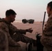 Two military personnel in desert camouflage uniforms are observed in the foreground, with one appearing to gesture or point toward a large military vehicle on a tarmac or airfield surface. A heavy military truck, consistent with a rocket artillery or logistics vehicle, is visible in the background on what appears to be a paved military installation. The scene is captured at dusk or dawn, giving the image a muted, low-light quality.