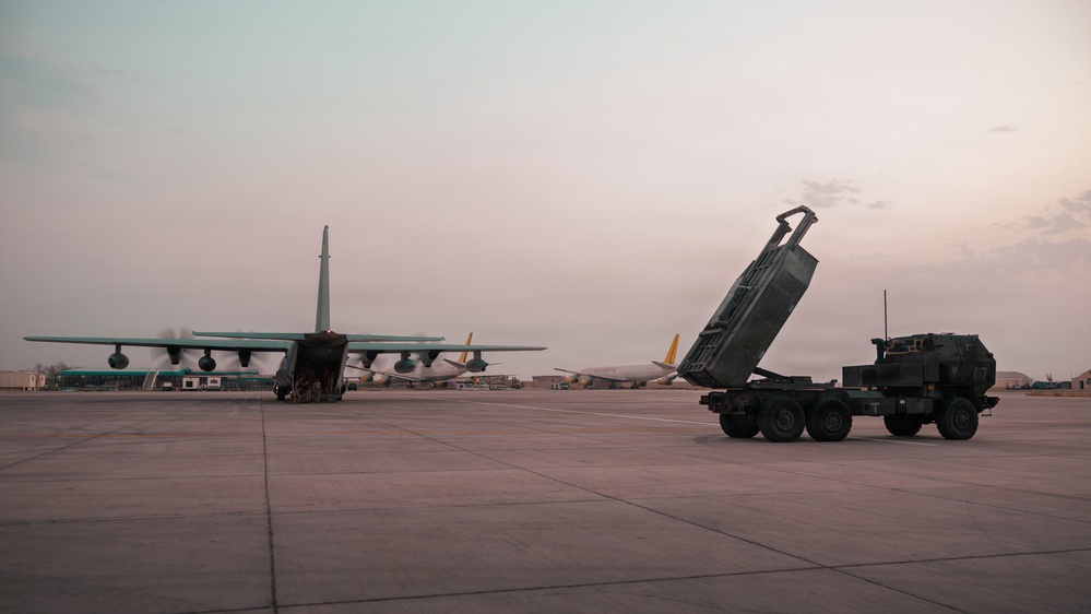 The image shows a military airfield at dusk or dawn with a large transport aircraft and a ground-based rocket artillery system positioned on the tarmac. A HIMARS (High Mobility Artillery Rocket System) launcher is visible with its pod elevated, suggesting recent deployment or preparation activities. Additional aircraft are visible in the background near what appears to be a terminal or hangar facility.