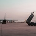 The image shows a military airfield at dusk or dawn with a large transport aircraft and a ground-based rocket artillery system positioned on the tarmac. A HIMARS (High Mobility Artillery Rocket System) launcher is visible with its pod elevated, suggesting recent deployment or preparation activities. Additional aircraft are visible in the background near what appears to be a terminal or hangar facility.