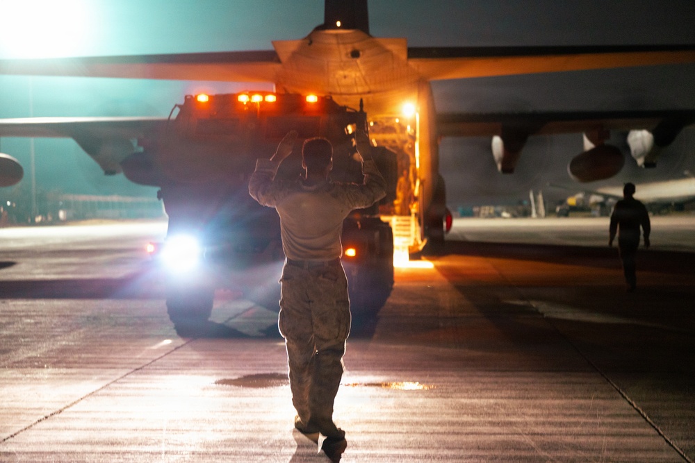 A nighttime military flight line operation showing a ground crew member marshaling a vehicle beneath a large military transport aircraft. The scene is illuminated by artificial lighting including vehicle headlights and aircraft lighting, creating a dramatic low-light atmosphere. A second personnel member is visible in the background walking near another aircraft.