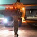 A nighttime military flight line operation showing a ground crew member marshaling a vehicle beneath a large military transport aircraft. The scene is illuminated by artificial lighting including vehicle headlights and aircraft lighting, creating a dramatic low-light atmosphere. A second personnel member is visible in the background walking near another aircraft.