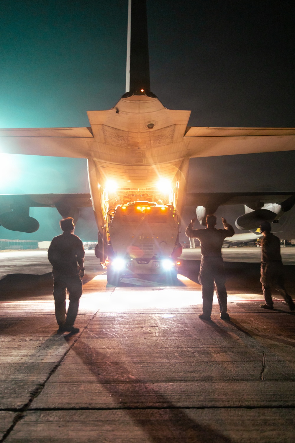 A nighttime military airfield operation showing a vehicle being loaded or unloaded from the rear cargo ramp of a large military transport aircraft. Ground crew personnel are silhouetted against the bright lights of the vehicle and aircraft interior, with at least one person using hand signals to guide the vehicle. The scene is illuminated by a combination of the aircraft's interior cargo lights, the vehicle's headlights, and ambient airfield lighting.