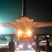 A nighttime military airfield operation showing a vehicle being loaded or unloaded from the rear cargo ramp of a large military transport aircraft. Ground crew personnel are silhouetted against the bright lights of the vehicle and aircraft interior, with at least one person using hand signals to guide the vehicle. The scene is illuminated by a combination of the aircraft's interior cargo lights, the vehicle's headlights, and ambient airfield lighting.