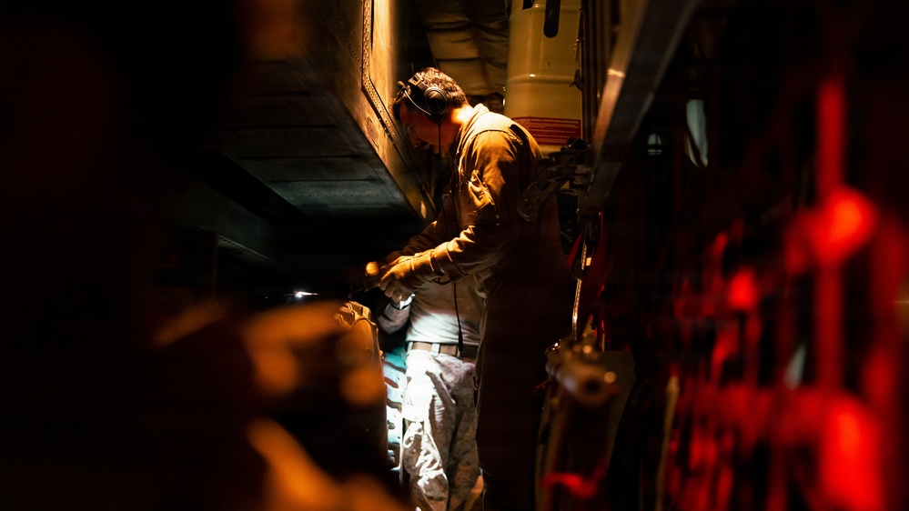 A person wearing aviation or military headset and flight suit is working inside what appears to be a military aircraft cargo hold or interior compartment. The scene is dramatically lit with a single light source illuminating the worker against a dark background, with red indicator lights visible on the right side. The individual appears to be performing maintenance, inspection, or equipment checks on aircraft systems.
