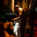 A person wearing aviation or military headset and flight suit is working inside what appears to be a military aircraft cargo hold or interior compartment. The scene is dramatically lit with a single light source illuminating the worker against a dark background, with red indicator lights visible on the right side. The individual appears to be performing maintenance, inspection, or equipment checks on aircraft systems.