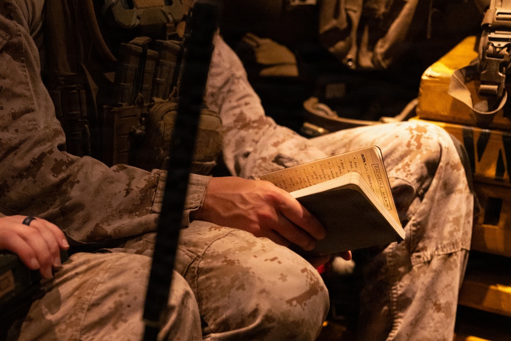 The image shows military personnel in desert camouflage uniforms seated in what appears to be a military vehicle or transport. One individual is holding and reading from a small notebook or field manual with handwritten notes visible on its pages. The scene is dimly lit with warm amber tones, suggesting low-light conditions inside a vehicle.