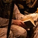 The image shows military personnel in desert camouflage uniforms seated in what appears to be a military vehicle or transport. One individual is holding and reading from a small notebook or field manual with handwritten notes visible on its pages. The scene is dimly lit with warm amber tones, suggesting low-light conditions inside a vehicle.
