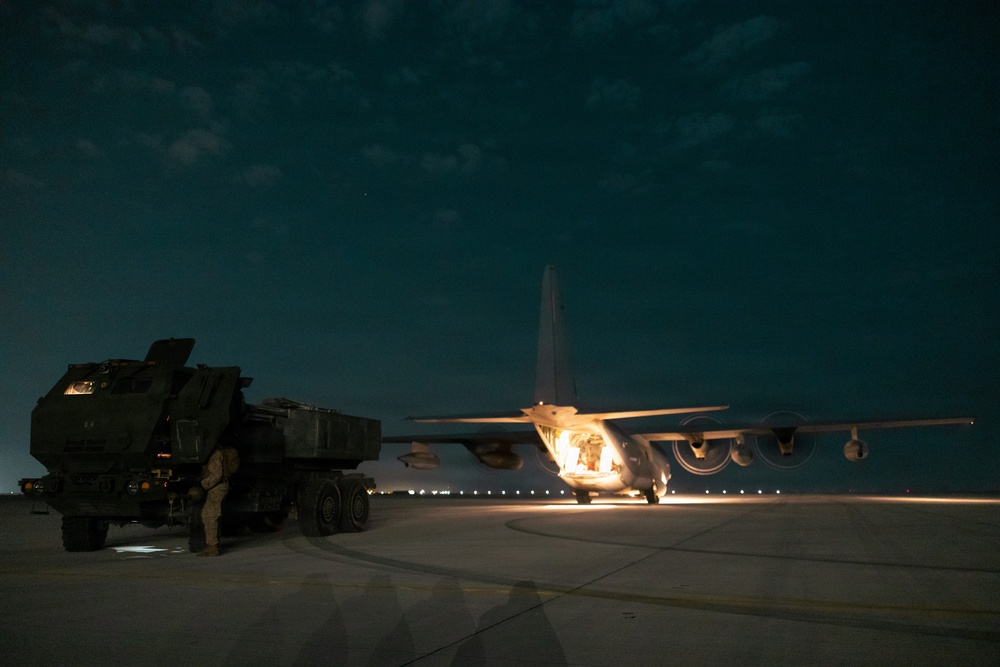 A nighttime military airfield operation showing a large transport aircraft with its rear cargo ramp open, positioned adjacent to a wheeled military rocket artillery system. The scene is illuminated by the aircraft's interior lighting and ground lights, suggesting a loading or unloading operation. Runway edge lights are visible in the background, confirming an active airfield environment.