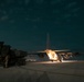A nighttime military airfield operation showing a large transport aircraft with its rear cargo ramp open, positioned adjacent to a wheeled military rocket artillery system. The scene is illuminated by the aircraft's interior lighting and ground lights, suggesting a loading or unloading operation. Runway edge lights are visible in the background, confirming an active airfield environment.