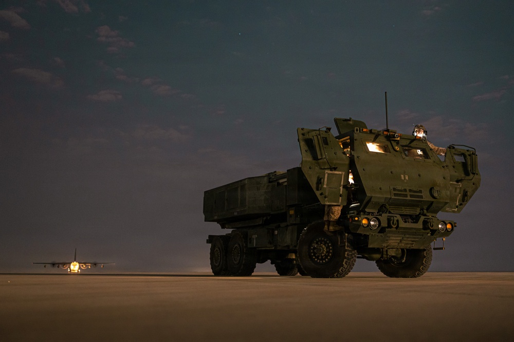 A nighttime military scene featuring a large olive-drab rocket artillery system, consistent with an M142 HIMARS or M270 MLRS launcher, positioned on what appears to be a tarmac or flat desert surface. At least one military personnel is visible in the cab area of the vehicle, illuminated by interior lighting. In the background to the left, a military transport aircraft — consistent with a C-130 Hercules — is visible on the ground with its lights on.