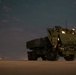 A nighttime military scene featuring a large olive-drab rocket artillery system, consistent with an M142 HIMARS or M270 MLRS launcher, positioned on what appears to be a tarmac or flat desert surface. At least one military personnel is visible in the cab area of the vehicle, illuminated by interior lighting. In the background to the left, a military transport aircraft — consistent with a C-130 Hercules — is visible on the ground with its lights on.