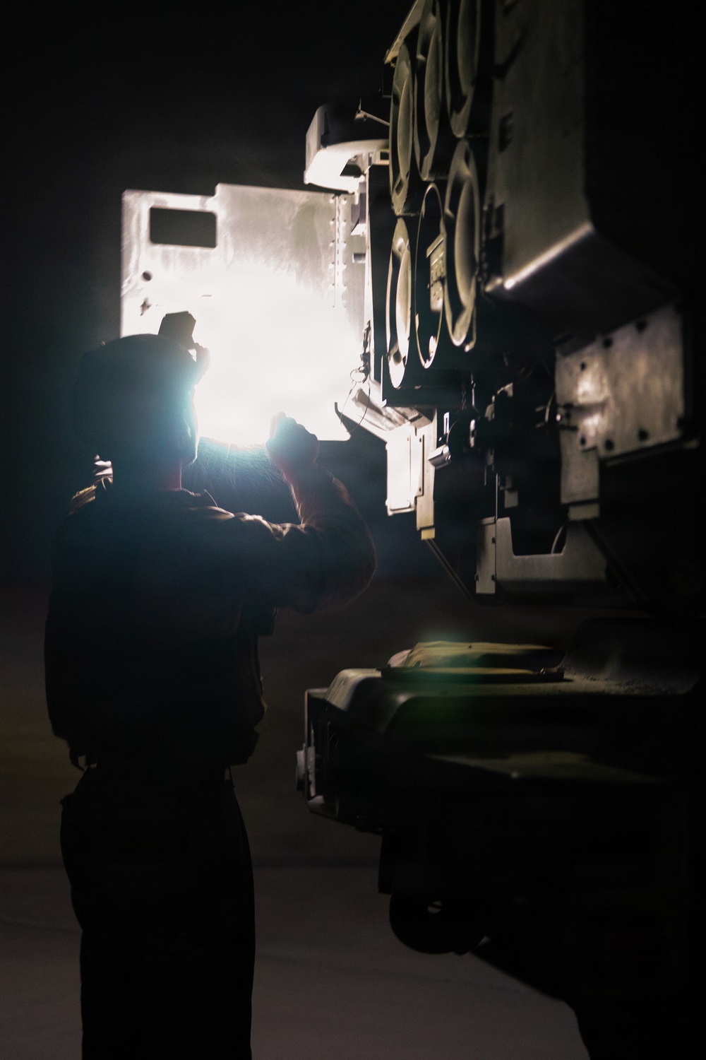 A silhouetted individual is working on or inspecting a large piece of military or heavy industrial equipment at night, using a bright light source to illuminate the work area. The scene is predominantly dark with the intense light creating a strong backlit effect that obscures facial and fine detail. The equipment visible appears to be a large armored or military vehicle with multiple circular components and modular panels.