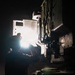A silhouetted individual is working on or inspecting a large piece of military or heavy industrial equipment at night, using a bright light source to illuminate the work area. The scene is predominantly dark with the intense light creating a strong backlit effect that obscures facial and fine detail. The equipment visible appears to be a large armored or military vehicle with multiple circular components and modular panels.