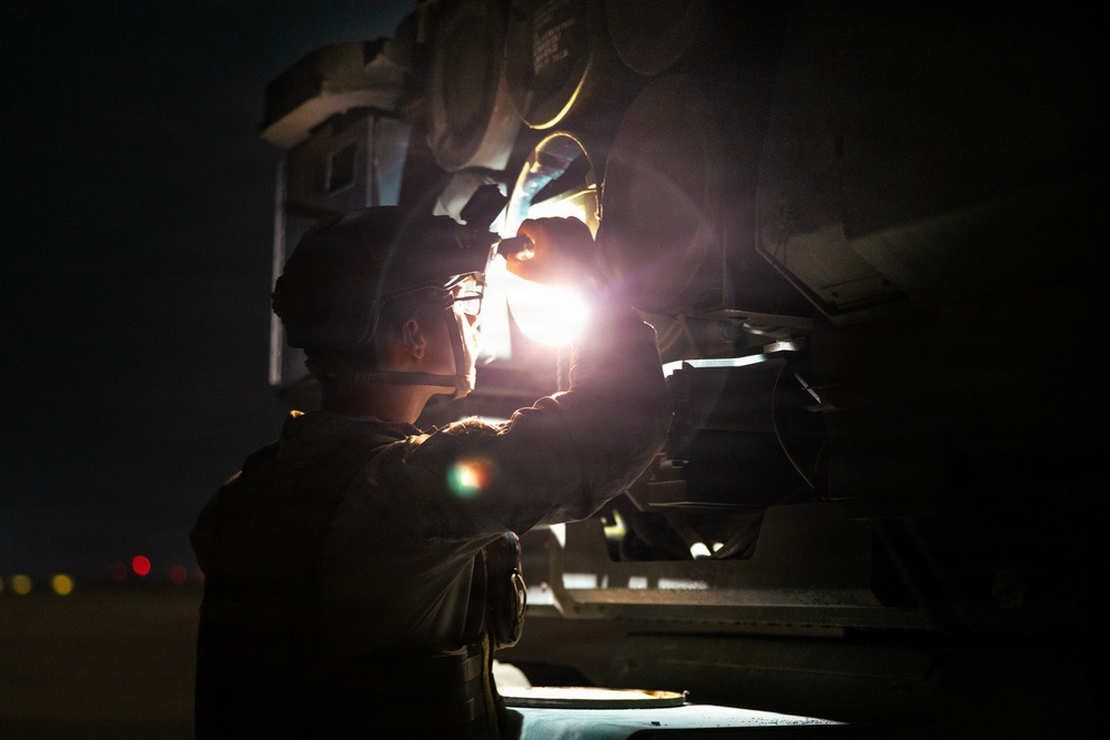 A military or aviation technician is performing a nighttime inspection or maintenance task on what appears to be a military aircraft or vehicle. The individual is using a handheld flashlight to illuminate the underside or mechanical components of the equipment. The scene is set on what appears to be a tarmac or airfield, with distant red and yellow lights visible in the background.