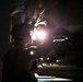 A military or aviation technician is performing a nighttime inspection or maintenance task on what appears to be a military aircraft or vehicle. The individual is using a handheld flashlight to illuminate the underside or mechanical components of the equipment. The scene is set on what appears to be a tarmac or airfield, with distant red and yellow lights visible in the background.