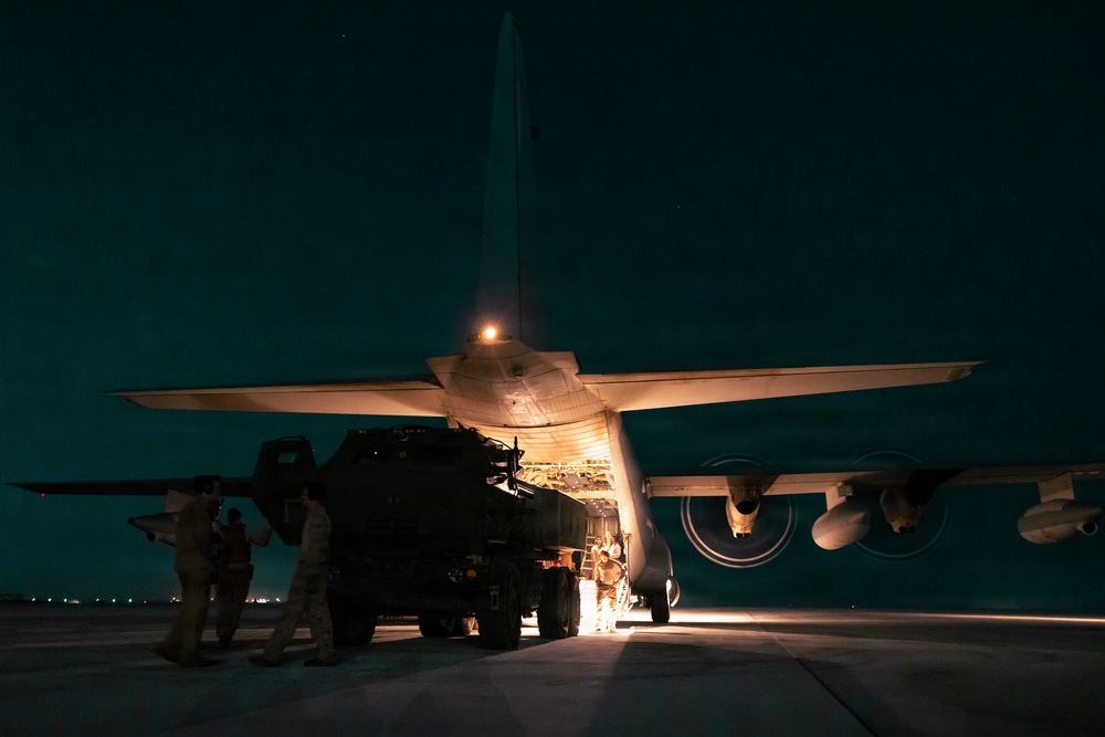 A nighttime military loading operation showing personnel loading a large military vehicle into the rear cargo ramp of a transport aircraft, likely a C-130 Hercules. The scene is illuminated by the aircraft's exterior lighting and interior cargo bay lights. Multiple military personnel are visible supervising and guiding the loading process.