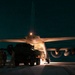 A nighttime military loading operation showing personnel loading a large military vehicle into the rear cargo ramp of a transport aircraft, likely a C-130 Hercules. The scene is illuminated by the aircraft's exterior lighting and interior cargo bay lights. Multiple military personnel are visible supervising and guiding the loading process.