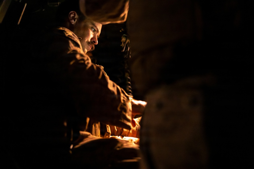 A man wearing military-style camouflage clothing is illuminated by a warm orange light source in an otherwise very dark environment. He appears to be focused on something in front of him, possibly working with equipment or materials at a low level. The dramatic chiaroscuro lighting creates a tense, atmospheric composition.