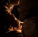 A man wearing military-style camouflage clothing is illuminated by a warm orange light source in an otherwise very dark environment. He appears to be focused on something in front of him, possibly working with equipment or materials at a low level. The dramatic chiaroscuro lighting creates a tense, atmospheric composition.