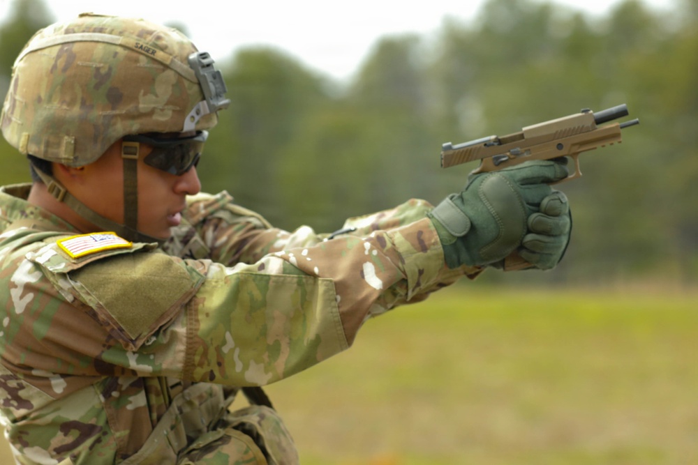 A U.S. Army soldier in MultiCam (OCP) uniform and ACH helmet is aiming a pistol in a two-handed grip at a firing range. The soldier is wearing tactical gloves, ballistic eyewear, and has a name tape reading 'SAGER' visible on the helmet. The firearm appears to be a SIG Sauer M17/M18 pistol, the U.S. Army's current standard-issue sidearm, identifiable by its coyote tan finish.