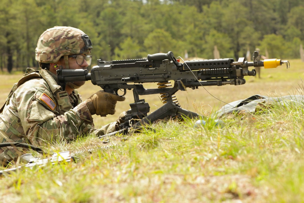 A U.S. Army soldier in MultiCam (OCP) camouflage uniform is in a prone firing position operating what appears to be an M240 machine gun mounted on a bipod. The soldier is positioned in a grassy field with a tree line visible in the background. A yellow muzzle cap/blank firing adapter is visible on the weapon, suggesting this is a training exercise.
