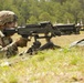A U.S. Army soldier in MultiCam (OCP) camouflage uniform is in a prone firing position operating what appears to be an M240 machine gun mounted on a bipod. The soldier is positioned in a grassy field with a tree line visible in the background. A yellow muzzle cap/blank firing adapter is visible on the weapon, suggesting this is a training exercise.