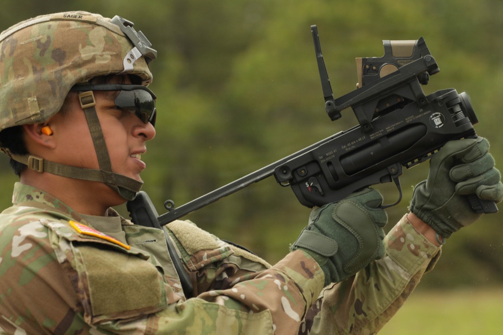 A U.S. Army soldier identified by the name tape 'SAGER' is handling what appears to be an XM25 Counter Defilade Target Engagement (CDTE) system or similar grenade launcher with an advanced optic/targeting system mounted on top. The soldier is wearing OCP (Operational Camouflage Pattern) uniform, tactical gloves, ballistic eyewear, and an ACH helmet with a device mounted on it. The image appears to be taken during a training or demonstration event in an outdoor setting.