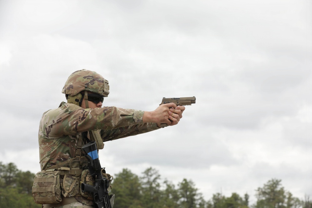 A soldier in full military combat gear is firing a pistol at a shooting range. The individual is wearing Operational Camouflage Pattern (OCP) uniform, a tactical helmet, and body armor with MOLLE webbing. The setting appears to be an outdoor military training range with overcast skies and treeline in the background.