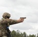 A soldier in full military combat gear is firing a pistol at a shooting range. The individual is wearing Operational Camouflage Pattern (OCP) uniform, a tactical helmet, and body armor with MOLLE webbing. The setting appears to be an outdoor military training range with overcast skies and treeline in the background.