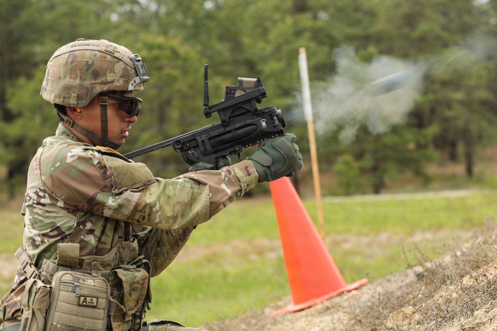 A U.S. Army soldier in MultiCam/OCP camouflage uniform is firing a standalone grenade launcher, likely an M320 or similar weapon system, during a training exercise. Smoke and a projectile are visibly captured mid-flight exiting the muzzle. The setting appears to be a military training range marked with an orange safety cone and wooden barrier.