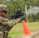 A U.S. Army soldier in MultiCam/OCP camouflage uniform is firing a standalone grenade launcher, likely an M320 or similar weapon system, during a training exercise. Smoke and a projectile are visibly captured mid-flight exiting the muzzle. The setting appears to be a military training range marked with an orange safety cone and wooden barrier.