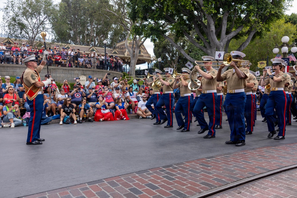 A U.S. Marine Corps marching band performs in what appears to be Disneyland's Town Square or Main Street U.S.A., likely during a Fourth of July or patriotic holiday celebration. Band members in dress blues with tan shirts march in formation while playing brass instruments, led by a drum major in full ceremonial uniform. A large crowd of spectators, many wearing red, white, and blue attire, watches from the sides and an elevated viewing area.