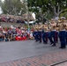 A U.S. Marine Corps marching band performs in what appears to be Disneyland's Town Square or Main Street U.S.A., likely during a Fourth of July or patriotic holiday celebration. Band members in dress blues with tan shirts march in formation while playing brass instruments, led by a drum major in full ceremonial uniform. A large crowd of spectators, many wearing red, white, and blue attire, watches from the sides and an elevated viewing area.
