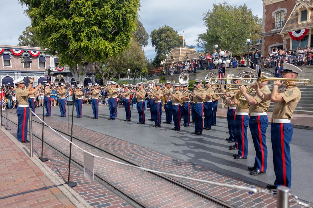 A U.S. Marine Corps band is performing in formation on a brick-paved street, likely at Disneyland's Main Street U.S.A., given the recognizable architecture and layout. The musicians are dressed in standard Marine Corps Service uniforms with tan shirts, blue trousers with red stripes, white belts, and white covers (caps). A large crowd of spectators watches from the sides and an elevated platform in the background, with red, white, and blue patriotic bunting decorating the buildings.