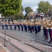 A U.S. Marine Corps band is performing in formation on a brick-paved street, likely at Disneyland's Main Street U.S.A., given the recognizable architecture and layout. The musicians are dressed in standard Marine Corps Service uniforms with tan shirts, blue trousers with red stripes, white belts, and white covers (caps). A large crowd of spectators watches from the sides and an elevated platform in the background, with red, white, and blue patriotic bunting decorating the buildings.