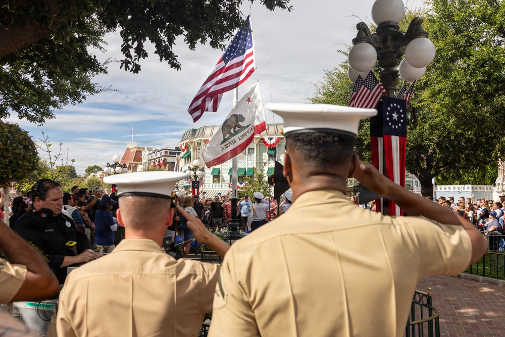 Two military personnel in tan dress uniforms and white covers (caps) are saluting during what appears to be a flag ceremony at Disneyland's Main Street, U.S.A. in California. Multiple flags are visible including the American flag, California Republic flag, and a Betsy Ross-style flag. A large crowd of spectators lines the street to observe the ceremony.