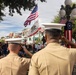 Two military personnel in tan dress uniforms and white covers (caps) are saluting during what appears to be a flag ceremony at Disneyland's Main Street, U.S.A. in California. Multiple flags are visible including the American flag, California Republic flag, and a Betsy Ross-style flag. A large crowd of spectators lines the street to observe the ceremony.