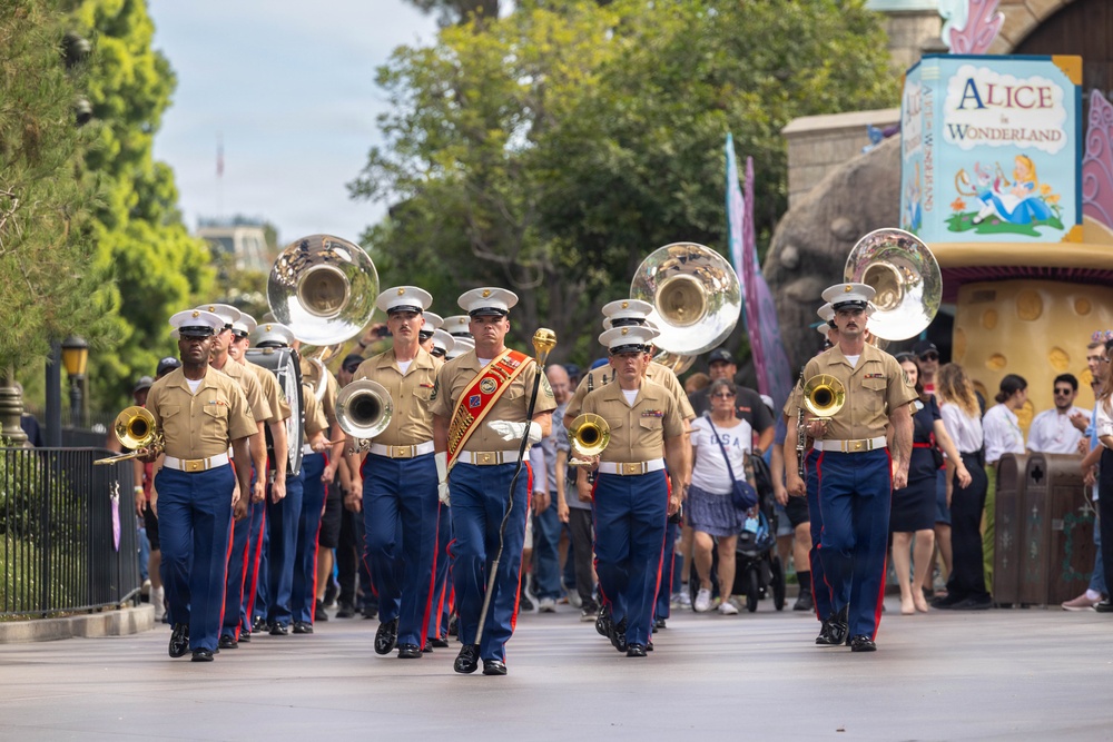 A U.S. Marine Corps marching band is performing a parade at what appears to be Disneyland, specifically near the Alice in Wonderland attraction. The band members are dressed in standard Marine Corps service uniforms with blue trousers featuring red stripes, and are carrying sousaphones and other brass instruments. A drum major leads the formation wearing a red ceremonial sash and carrying a mace.