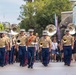 A U.S. Marine Corps marching band is performing a parade at what appears to be Disneyland, specifically near the Alice in Wonderland attraction. The band members are dressed in standard Marine Corps service uniforms with blue trousers featuring red stripes, and are carrying sousaphones and other brass instruments. A drum major leads the formation wearing a red ceremonial sash and carrying a mace.