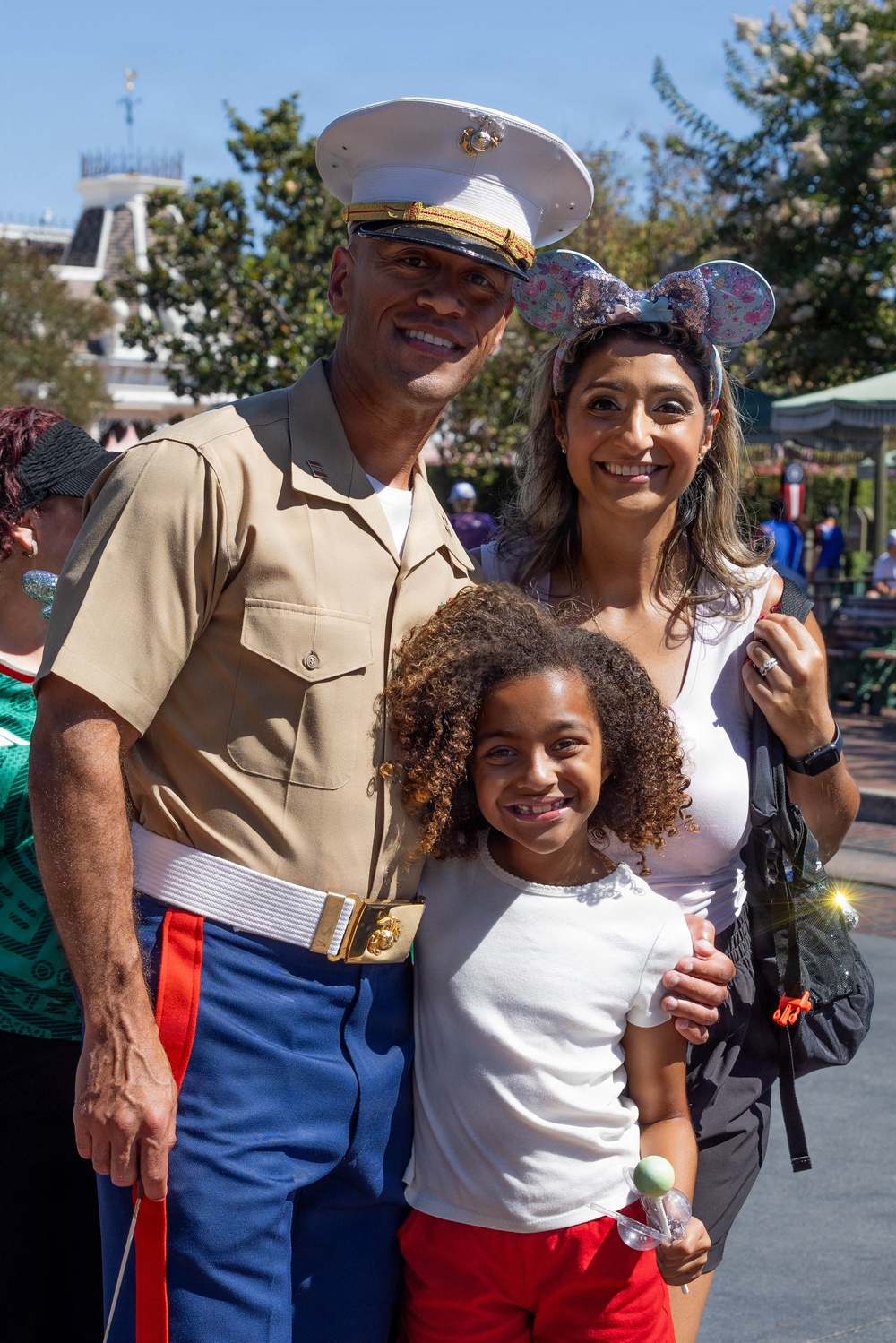 A family of three poses for a photo at what appears to be Disneyland, identifiable by the park's Main Street architecture in the background. The adult male is wearing a United States Marine Corps dress uniform (Service Charlie), while the woman wears Mickey Mouse ears headband and the young girl holds a small toy or lollipop. The group is smiling and appears to be enjoying a family outing at the theme park.