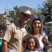 A family of three poses for a photo at what appears to be Disneyland, identifiable by the park's Main Street architecture in the background. The adult male is wearing a United States Marine Corps dress uniform (Service Charlie), while the woman wears Mickey Mouse ears headband and the young girl holds a small toy or lollipop. The group is smiling and appears to be enjoying a family outing at the theme park.