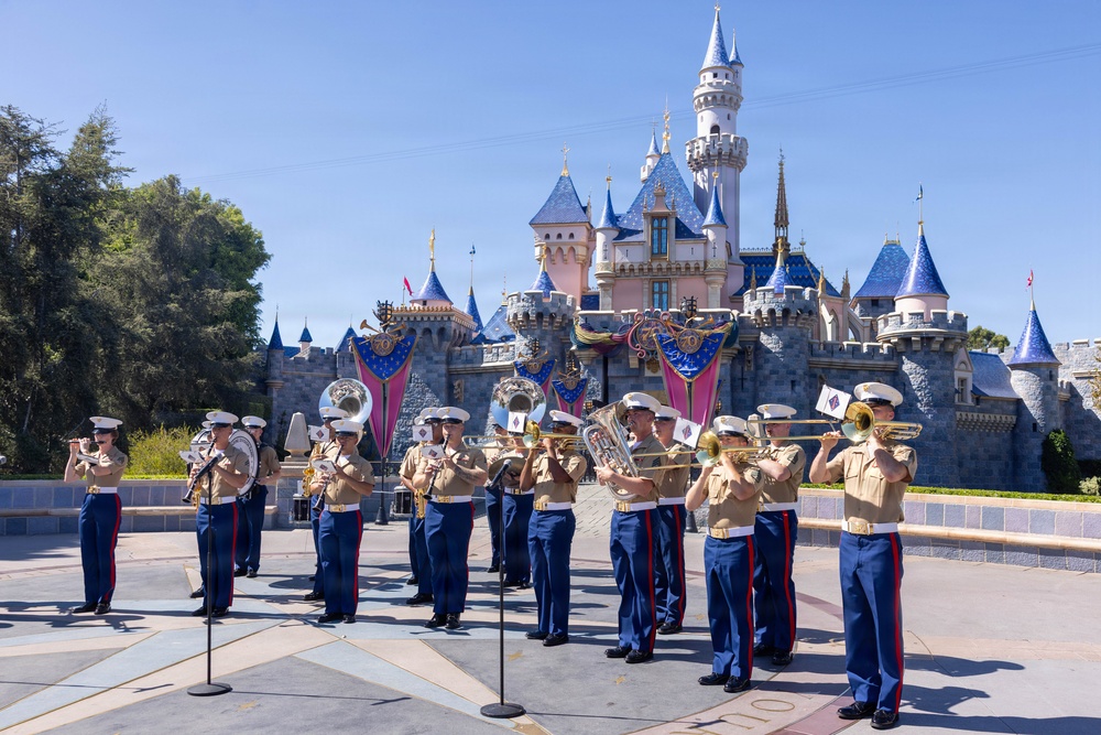 A U.S. Marine Corps marching band performs in front of Sleeping Beauty Castle at Disneyland. The musicians are dressed in formal Marine dress uniforms featuring tan blouses, blue trousers with red stripes, and white peaked caps. The performance appears to be an official military band event at the theme park, likely during a special ceremony or celebration.
