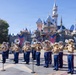 A U.S. Marine Corps marching band performs in front of Sleeping Beauty Castle at Disneyland. The musicians are dressed in formal Marine dress uniforms featuring tan blouses, blue trousers with red stripes, and white peaked caps. The performance appears to be an official military band event at the theme park, likely during a special ceremony or celebration.