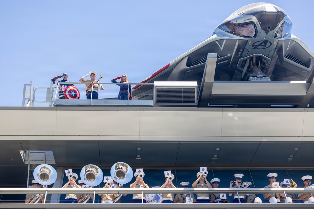 The image shows a Marvel-themed event, likely at a Disney theme park or cruise ship, featuring costumed characters including Captain America on an upper deck alongside what appears to be a military band member and another character. A large Avengers-branded aircraft or quinjet prop is prominently displayed on the upper level. Below, a marching band in white uniforms performs with brass instruments including tubas and trumpets.